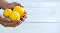 Person with medium skin tone holding three fresh yellow lemons in cupped hands against white wooden background, suitable for food, health, and culinary content.