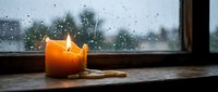 Burning orange candle on wooden windowsill during rainy day creates cozy atmospheric mood for relaxation and meditation indoors.