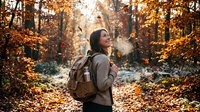 Young Asian woman hiker breathing cold air in autumn forest with golden leaves, wearing backpack and warm sweater during outdoor adventure trek.
