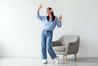 Young woman dancing with raised hands in casual blue outfit at home interior with modern armchair on white background.