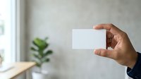 Young adult male with medium brown skin tone holding blank white business card in professional setting with natural light and potted plant background.