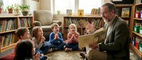 Elderly Caucasian grandfather reading storybook to diverse group of young children sitting on library floor surrounded by bookshelves and plants.