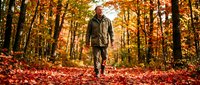 Middle-aged man with light skin tone wearing olive jacket jogging on autumn forest path covered in fallen leaves during fall season.