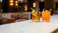 Two craft cocktails with garnishes on marble bar counter in upscale restaurant with warm ambient lighting for hospitality and beverage marketing.