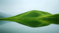 Serene green rolling hills reflected in still lake waters create mirror symmetry against misty mountain backdrop, capturing tranquil natural landscape.