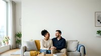 Young couple with dark hair sitting on gray sofa in bright living room with potted plants, having conversation near window with natural light for home lifestyle and relationship content.