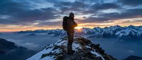 Adventurous hiker with backpack standing on snowy mountain peak at dramatic sunrise, overlooking alpine landscape with snow-capped peaks and colorful sky.