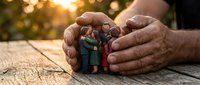 Protective hands cupping miniature family figures on wooden surface with warm sunlight, symbolizing care, security and family protection concept.