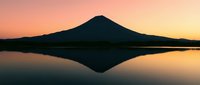 Silhouette of a snow-capped volcanic mountain reflected in still lake water during golden hour sunrise or sunset with warm orange and pink sky.