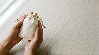 Person with medium skin tone holding small beige cotton drawstring pouch on neutral linen surface for product packaging and gift wrapping concepts.