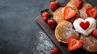 Fresh pancakes with ripe strawberries, whipped cream heart decoration and powdered sugar on dark wooden board against black background, copy space.