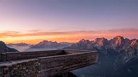 Wooden railing overlooks dramatic mountain peaks at sunrise with golden and pink sky, alpine valley below, scenic landscape for travel and nature projects.