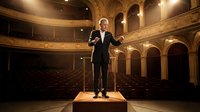 Middle-aged Caucasian male conductor in formal black tuxedo performing on stage in elegant classical theater with ornate golden architecture and tiered seating.