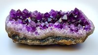 Raw amethyst crystal cluster with deep purple crystalline formations growing from natural stone base, displaying geometric shapes and varying color intensity.
