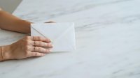 Woman with medium skin tone holding blank white envelope on marble surface, suitable for business correspondence, stationery mockups, and professional communication concepts.