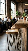 Wooden bar stools with metal legs at rustic counter in industrial loft cafe with brick walls, large windows, and blurred customers socializing in background for restaurant and hospitality design.