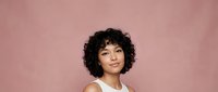 Young mixed-race woman with curly black hair wearing white tank top against pink background, confident expression for lifestyle and fashion content.