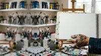 Artist holding open spiral notebook with blank page in creative studio workspace surrounded by art supplies and paintbrushes.