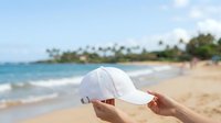 Young woman holding white baseball cap at tropical beach with palm trees and ocean waves, suitable for summer vacation, travel, and lifestyle marketing campaigns.