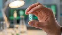 Hand holding white pills or tablets between fingers in modern laboratory or medical facility with blurred equipment background for healthcare concepts.