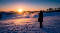 Person standing alone in snowy winter landscape at sunrise, watching golden sun over frozen field with bare trees. Solitude and contemplation concept for wellness and nature themes.