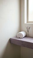 Rolled white towel and dried lavender flowers on rustic wooden shelf by window in minimalist spa bathroom setting for wellness and relaxation design.