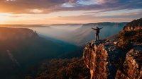 Hiker with raised arms celebrating success on mountain cliff edge at golden sunrise overlooking misty valley canyon landscape adventure achievement concept.