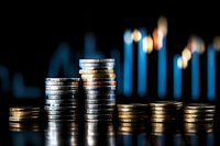 Stacks of silver and gold coins arranged in ascending and descending order against dark background with blurred financial chart showing market trends.