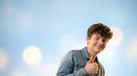 Young Caucasian man with curly hair giving thumbs up gesture against blue sky with clouds, expressing positivity and success in casual denim shirt.