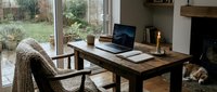 Cozy home office with rustic wooden desk, laptop, candle, and sleeping dog by fireplace, overlooking rainy garden through large window.