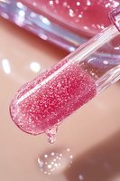 Pink glittery lip gloss applicator with shimmering sparkles and droplet falling, macro shot against soft blurred background showing luxurious cosmetic texture.