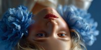 Young blonde woman with blue eyes lying upside down surrounded by vibrant blue peony flowers against soft background for beauty and wellness concepts.