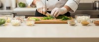 Professional chef in white uniform chopping fresh green herbs on wooden cutting board in modern commercial kitchen with ingredients and spices.