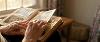Elderly hands turning pages of vintage book in warm sunlight streaming through window creating peaceful reading atmosphere for literature and education concepts.