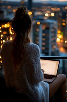Young woman in white knit sweater working on laptop at night, city lights bokeh in background, sitting on balcony with fairy lights creating cozy atmosphere.