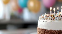 Festive birthday cake with white frosting and chocolate sprinkles, lit candles glowing against blurred colorful balloon background, close-up view of celebration dessert.