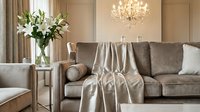 Elegant living room with gray upholstered sofa, draped throw blanket, white lilies in glass vase, crystal chandelier, and neutral interior design for home decor inspiration.
