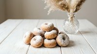 Fresh homemade donuts dusted with powdered sugar arranged on rustic white wooden table with dried pampas grass in glass vase creating cozy bakery atmosphere.