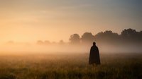 Silhouette of person standing in misty field at golden hour sunrise with warm atmospheric light and peaceful countryside landscape for meditation concepts.