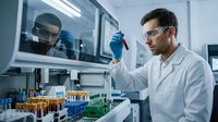 Male scientist in white lab coat and safety glasses examining test tube in modern laboratory setting with equipment and samples on workbench for research and development.