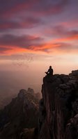 Silhouetted male figure sitting on rocky mountain cliff edge at sunset with flock of birds flying overhead, dramatic orange and purple sky, misty valley below, contemplative mood for meditation and freedom themes.