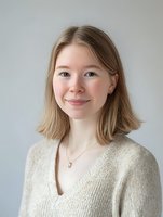 Young Caucasian woman with blonde shoulder-length hair smiling warmly at camera, wearing beige knit sweater against light gray background. Natural daylight portrait.
