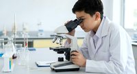 Young Asian male scientist in white lab coat examining samples through microscope in modern laboratory for research and medical analysis.