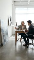 Mature Caucasian businesswoman consulting with Asian male client in modern office space with artwork and natural light for professional services.