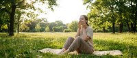 Woman in casual beige clothing sitting on grass in sunny park, relaxing outdoors among trees and green meadow for lifestyle and wellness content.