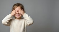Young girl covering eyes with hands in playful peek-a-boo gesture against neutral background for childhood development and parenting concepts.