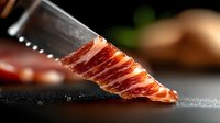 Thinly sliced premium cured meat on dark surface with knife blade in macro shot showing marbled texture and rich red color for gourmet food photography.