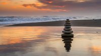 Zen stone stack balanced on wet sand reflects in ocean water during golden sunset with waves and colorful sky for meditation and mindfulness concepts.