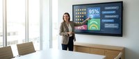 Professional businesswoman presenting analytics dashboard on large wall-mounted display in modern conference room with data visualization charts.
