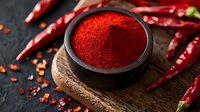 Vibrant red chili powder in ceramic bowl on rustic wooden board with dried red chili peppers scattered around on dark background, macro food photography.